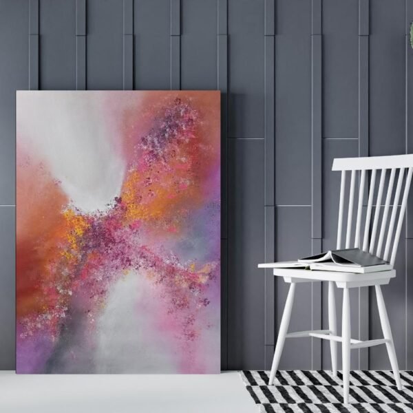 Pink and orange abstract canvas art leaning against a dark paneled wall beside a white wooden chair with books on it.